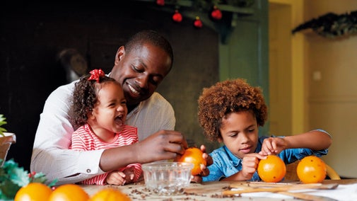 A father and two happy children make Christmas decorations, putting cloves into oranges. There are Christmas wreaths in the background